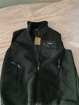 Patagonia väst black - Säljer en svart fleeceväst från Patagonia i storlek M. Västen har mjukt teddyfoder, dragkedja framtill, två sidofickor med dragkedja och en bröstficka med dragkedja. Perfekt för lager-på-lager och riktigt skön att ha på sig.  snygg helsvart Patagonia-väst 🖤 Klassisk modell som passar perfekt till både vardag och streetwear.  • Färg: Svart • Storlek: M • Skick: ny med prislapp  • Äkta Patagonia • Säljs då den är för stor  Skickar snabbt och packar väl 📦 Hör gärna av dig vid intresse