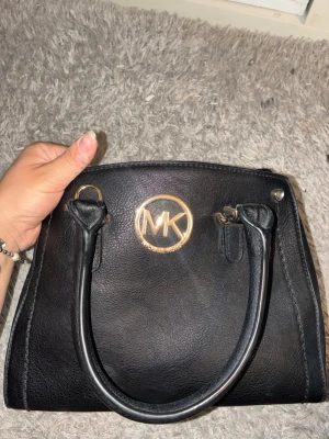 Svart handväska med gulddetaljer - Snygg svart handväska med ett stort guldfärgat emblem framtill. Klassisk fyrkantig form och diskreta sömmar längs kanterna. Perfekt för dig som gillar stilrena accessoarer.