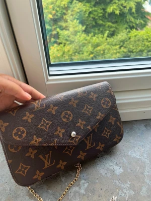 väska  - Snygg brun handväska med klassiskt monogrammönster i guld och brunt. Väskan har en elegant kuvertformad design och en tunn guldfärgad kedja som axelrem. Tillverkad i tåligt canvasmaterial med tryckknapp framtill.