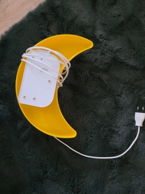 Gul månlampa med sladd - Säljer en dekorativ vägglampa i form av en gul måne. Lampan är gjord av plast och har en vit sladd med stickkontakt för enkel anslutning till eluttag. Perfekt som mysbelysning i sovrum eller barnrum.