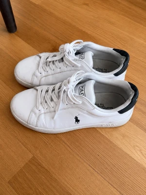 Vita Polo Ralph Lauren sneakers - Se defekter på bildena! Snygga vita sneakers från Polo Ralph Lauren med klassisk låg profil och svart detalj vid hälen. Skorna har snörning, diskret logga på sidan och är tillverkade i slätt läder. Perfekta för en clean och stilren look.