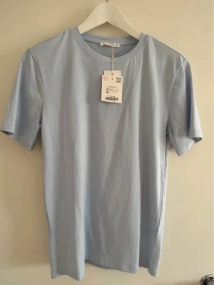 Ljusblå t-shirt från Zara  - En stilren ljusblå t-shirt med rund hals och korta ärmar. Tillverkad i mjuk bomull för en skön känsla. Perfekt för dig som gillar en enkel och clean look. Passar till det mesta och är lätt att matcha med andra plagg. En snygg T-shirt från Zara i ljusblå färg Skick: Nyskick Storlek S  Har endast tagit bort prislappen och aldrig använts! Helt ny!  