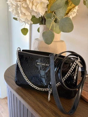 Svart lackad handväska Michael Kors - Snygg svart handväska från Michael Kors i glansigt lackmaterial med präglad MK-logga. Väskan har både en silverfärgad kedjerem och en justerbar axelrem i läder. Perfekt storlek för det viktigaste och stängs med dragkedja.
