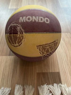 Gul och vinröd basketboll Mondo - Säljer en basketboll från Mondo i storlek 5. Bollen är gul och vinröd med tryck av basketkorg och spelare. Perfekt för träning eller spontanspel med vänner. Greppvänlig yta för bättre bollkontroll.