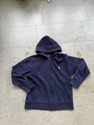 Mörkblå hoodie med dragkedja Polo - Mörkblå hoodie från Polo Ralph Lauren med vit broderad logga på bröstet. Hoodien har dragkedja framtill, två fickor och justerbar huva med snören. Tillverkad i mjuk bomull och har långa ärmar med muddar.