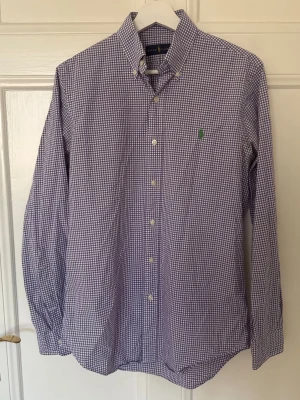 Lila rutig skjorta från Ralph Lauren - Snygg lila och vit rutig skjorta från Ralph Lauren med klassisk krage och knappar framtill. Skjortan har lång ärm och en liten grön logga broderad på bröstet. Perfekt för en stilren look.