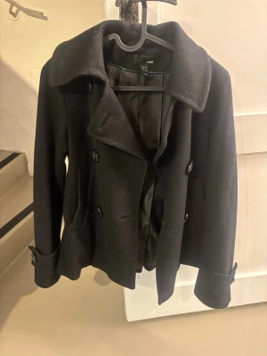 Svart kappa H&M - Stilren svart kort kappa från H&M med dubbelknäppning och bred krage. Rocken har två fickor framtill och dekorativa knappar vid ärmsluten. Tillverkad i ullblandning och helfodrad för extra komfort. Perfekt för kyliga dagar.