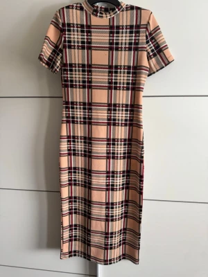 Beige rutig klänning med kort ärm - Trendig beige klänning med rutigt mönster i svart, rött och vitt. Modellen har korta ärmar, rund halsringning och rak passform. Perfekt för dig som gillar klassisk men ändå modern stil.