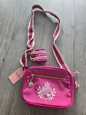Rosa axelväska från Juicy Couture - Snygg rosa axelväska från Juicy Couture med justerbar randig axelrem i rosa och vitt. Väskan har guldfärgad dragkedja, hjärtberlock och broderad logga framtill. Perfekt för dig som vill ha en trendig och praktisk väska med coola detaljer.