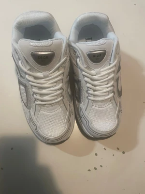 Vita Di0r sneakers med grå detaljer - Snygga vita sneakers från Di0r med grå och silverfärgade detaljer. Skorna har en chunky sula, meshpaneler och klassisk snörning. Perfekta för dig som vill ha en exklusiv och trendig look. Märkeslogga på plösen och sidan.