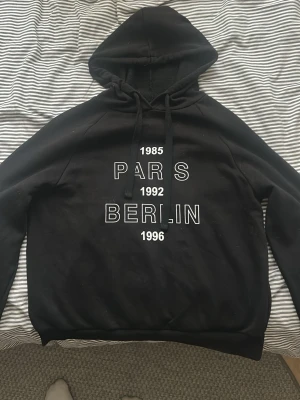 Svart hoodie med tryck Paris Berlin - Svart hoodie med huva och dragsnören. Tryck på framsidan med texten '1985 PARIS 1992 BERLIN 1996' i vitt. Klassisk passform med ribbade muddar vid ärmslut och nederkant. Perfekt för en avslappnad streetwear-look. Jättebillig och Prisvärd! Kan diskutera pris.