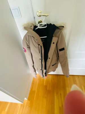 Beige pufferjacka med luva Canada Goose - Snygg beige pufferjacka från  Canada Goose med luva och fuskpälsdetalj. Jackan har ribbade muddar, två stora fickor framtill och en patch på ärmen. Perfekt för kalla vinterdagar och har en klassisk, tidlös look. Storlek XXS men passar en liten S och XS. Fint skick.