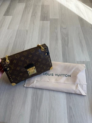 Louis Vuitton monogram handväska - Snygg handväska från Louis Vuitton i klassiskt brunt monogrammönster. Väskan har guldfärgade metalldetaljer, kedjerem och dekorativt lås framtill. Tillverkad i tåligt läder med svarta detaljer och kommer med dustbag.