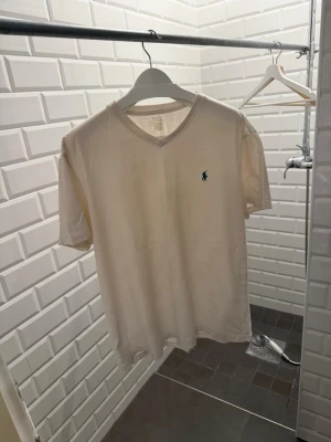 Creme förgad t-shirt från Polo Ralph Lauren - Creme t-shirt med v-ringning från Polo Ralph Lauren. Klassisk passform och korta ärmar, med den ikoniska blå loggan broderad på bröstet. Perfekt basic-plagg i mjuk bomull som funkar till allt.
