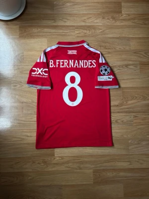 Man United hemma matchtröja B. Fernandes #8 | storlek: M - Säljer en röd Manchester United matchtröja med vita detaljer och nummer 8 B. Fernandes på ryggen. Tröjan har klubbmärke, Adidas-logga och sponsorer på bröstet samt Champions League-märke på ärmen. Klassisk krage och korta ärmar. Perfekt för fotbollsfans!