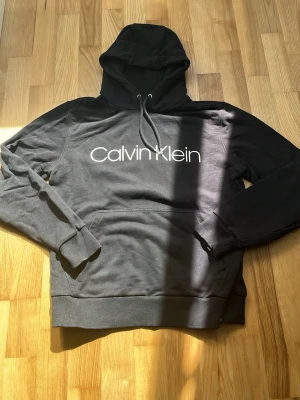 Svart hoodie från Calvin Klein - Snygg svart hoodie från Calvin Klein med stor vit logga på bröstet. Klassisk modell med huva och dragsnören samt magficka framtill. Tillverkad i mjukt material som känns skönt mot huden. Perfekt för en avslappnad och trendig look.