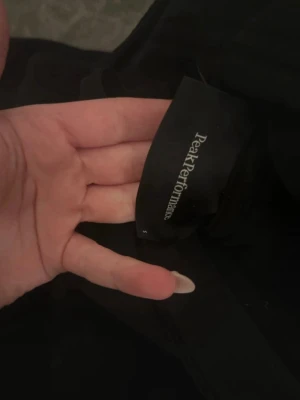 Svart hoodie från Peak Performance - Snygg svart hoodie från Peak Performance med vit logga på bröstet. Klassisk modell med huva och dragsko samt en stor magficka framtill. Tillverkad i mjukt material som passar perfekt till chill eller streetwear.