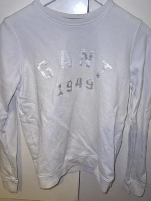 Vit sweatshirt från GANT - Klassisk vit sweatshirt från GANT med tryckt logga och årtalet 1949 i silver på bröstet. Tröjan har rund halsringning, långa ärmar och ribbade muddar vid ärmslut och nederkant. Perfekt för en clean och stilren look.