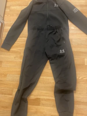Svart overall från Under Armour - Säljer en svart overall från Under Armour med dragkedja framtill, huva och resår i midjan. Overallen har tryckta loggor på bröstet, benet och ärmen samt muddar vid bensluten. Perfekt för träning eller chill. Materialet känns mjukt och stretchigt.