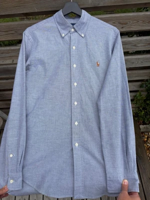 Blå skjorta från Ralph Lauren - Klassisk blå skjorta från Ralph Lauren med button-down krage och broderad logga på bröstet. Skjortan har långa ärmar och vita knappar framtill. Tillverkad i mjuk bomull som känns skön mot huden. Perfekt för en stilren och avslappnad look.