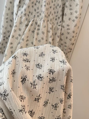 Smockad maxiklänning  - Romantisk långklänning i vit bomull med svart blommigt mönster. Klänningen har långa puffärmar med smock vid ärmslut, rund halsringning och lätt rynkad midja för en feminin siluett. Perfekt för alla tillfällen.