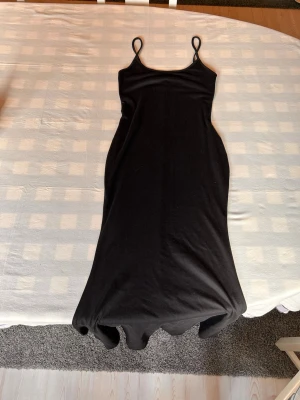 Svart långklänning med smala band - En enkel och stilren svart långklänning med smala axelband och rak passform. Klänningen har en minimalistisk design och är perfekt för dig som gillar en clean look. Materialet känns mjukt och följsamt mot kroppen.