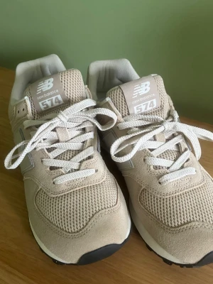 Beige New Balance 574 sneakers - Stilrena beige New Balance 574 sneakers med ovandel i mocka och mesh. Skorna har vita snören, klassisk N-logga på sidan och en bekväm sula i vitt och svart. Perfekta för dig som gillar en clean och sportig look.