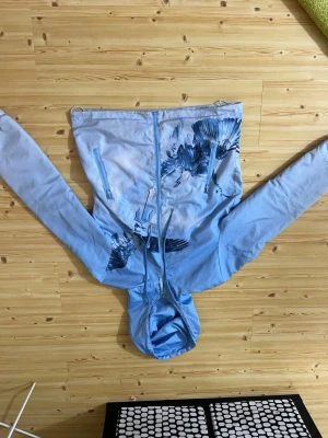 Ljusblå vindjacka med fågeltryck - Snygg ljusblå vindjacka med huva och dragkedja framtill. Jackan har coolt fågeltryck på bröstet och sidorna samt två dragkedjeförsedda fickor. Perfekt för blåsiga dagar och enkel att matcha med jeans eller träningsbyxor. Lite smutsig fast tvättar bort de innan jag skickar den 
