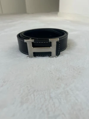 Harmes bälte - Snyggt svart skärp med kontrastsömmar och ett stort, silverfärgat H-format spänne. Skärpet har en stilren design och är tillverkat i skinn, vilket ger en exklusiv känsla. Perfekt accessoar för att lyfta din outfit.