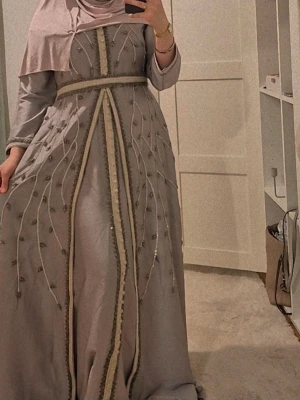 Lång ljuslila klänning med broderi - Elegant långklänning i ljuslila med broderade detaljer och pärlor längs hela klänningen. Klänningen har lång ärm och markerad midja med dekorativa band. Perfekt för speciella tillfällen och ger en lyxig känsla.