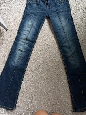 Mörkblå bootcut jeans - Säljer ett par mörkblå bootcut jeans med snygga slitningar framtill. Jeansen har klassisk femficksdesign och kontrastsömmar. Perfekta för dig som gillar en avslappnad och trendig look.