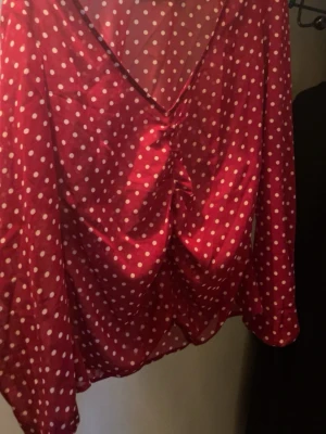 Röd prickig blus från NA-KD - Snygg röd blus från NA-KD med vita prickar och v-ringning. Blusen har långa ärmar och är gjord i ett lätt, glansigt material som ger en cool och trendig look. Perfekt för dig som gillar färg och mönster!
