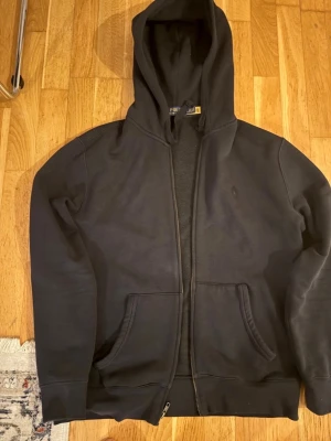 Svart hoodie med dragkedja - En svart hoodie med dragkedja framtill och huva. Hoodien har två stora fickor på magen och långa ärmar. Perfekt för en avslappnad och stilren look.