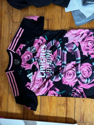 Svart och rosa Real Madrid tröja - Unik svart och rosa Real Madrid matchtröja från Adidas med stora rosa rosor och en orm på framsidan. Tröjan har korta ärmar, rosa detaljer på axlar och ärmslut samt klubbmärke och sponsortryck på bröstet. Tillverkad i lätt och ventilerande material.