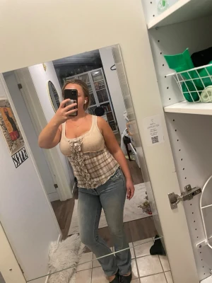 Beige rutig topp med volangdetaljer - Säljer en beige och ljusbrun rutig topp med breda axelband och volangdetaljer framtill. Toppen har en figurnära passform och är gjord i ett mjukt, stretchigt material. Perfekt för dig som gillar en söt och trendig stil.