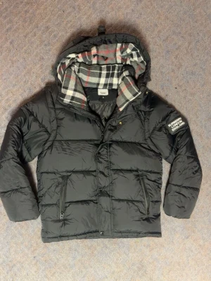 Svart pufferjacka med huva - Snygg svart pufferjacka med rutigt foder i huvan och dragkedjefickor. Jackan har en patch på ärmen med texten 'BURBERRY LONDON ENGLAND' och en mysig huva med grått, vitt och rött rutmönster. Perfekt för kalla dagar. Priset är inte hugget i sten