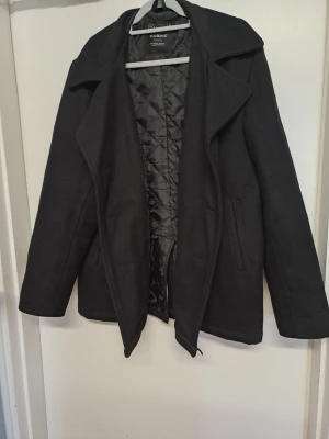 Klassisk svart rock  - Stilren svart kappa med bred krage och quiltat innerfoder. Kappan har två sidofickor och rak passform, perfekt för kyliga dagar. designen är tidlös och enkel.katt finns i hemmet