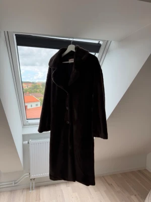Long Dark Brown Faux Fur Coat - A long, dark brown faux fur coat with a wide collar and button closure. The coat appears to be mid-calf length and has a relaxed, slightly oversized fit. It is shown both hanging on a hanger in front of a window and worn by a person in front of a mirror.