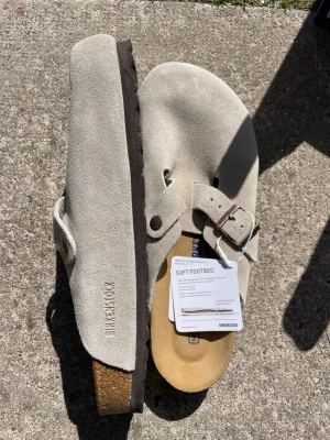 Beige mockatofflor från Birkenstock - Säljer ett par snygga beige tofflor från Birkenstock i mjuk mocka med justerbart spänne i metall. Tofflorna har en klassisk korksula och mjuk fotbädd för extra komfort. Perfekta för dig som vill ha både stil och bekvämlighet. OG Kartong  ingår