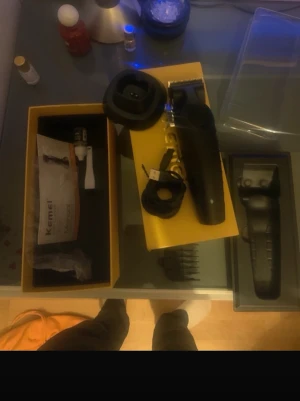 Svart hårtrimmer med tillbehör - Säljer en svart hårtrimmer från Kemei med flera tillbehör som laddkabel, rengöringsborste och olika kammar. Allt kommer i originalförpackning med gul och svart design. Perfekt för styling och trimning av hår och skägg.