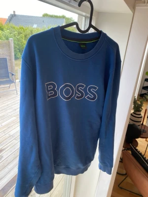 Blå sweatshirt från BOSS - Snygg blå sweatshirt från BOSS med rund halsringning och vit broderad BOSS-logga på bröstet. Tröjan har ribbade muddar vid ärmslut och nederkant. Perfekt för en avslappnad och trendig look.