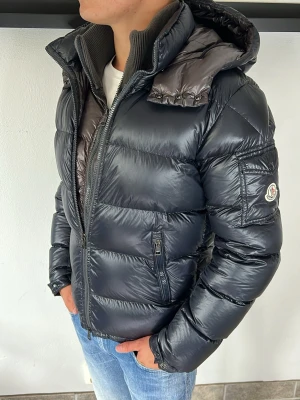 Moncler Jacka  - Snygg svart dunjacka från Moncler med glansig finish och vadderad design. Jackan har en stor huva, två framfickor med dragkedja och Moncler-logga på ärmen. Perfekt för kalla vinterdagar och riktigt stilren look.