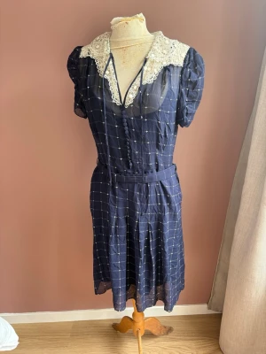 Marinblå vintageklänning med spetskrage Ralph Lauren stl 6 - Söt marinblå kortklänning stl 6  med rutigt mönster och dekorativa knappar framtill. Klänningen har en stor vit spetskrage och korta puffärmar. Bälte i midjan och lätt transparent tyg ger en retrovibe. Perfekt för dig som gillar unika vintageplagg. Helt ny med lapparna kvar. 