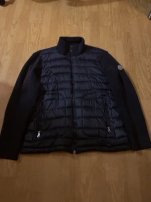 Svart pufferjacka från Moncler - Snygg svart pufferjacka från Moncler med quiltad front och stickade ärmar. Jackan har hög krage, dragkedja framtill och två fickor med dragkedja. Insidan har Monclers klassiska serietidningspatch. Perfekt för kalla dagar.