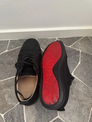Svarta låga sneakers med röd sula - Snygga svarta sneakers med lågt skaft och klassisk snörning. Ovandelen är i textil och mockaliknande material, medan sulan är svart med en cool röd undersida. Insidan är ljusbeige för en schysst kontrast. Perfekta för dig som gillar stilrena men ändå unika skor.