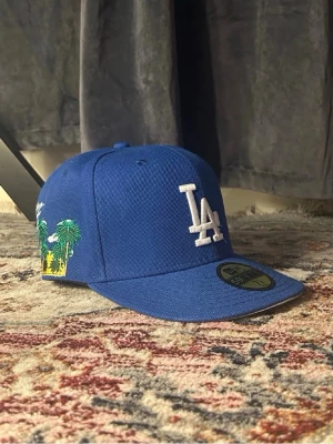 Blå LA keps med broderade palmer - Snygg blå keps med LA-logga framtill och broderade palmer på sidan. Kepsen har rak skärm och klassisk snapback-modell. Tillverkad i ett tåligt tyg och har New Era-logga broderad på sidan.