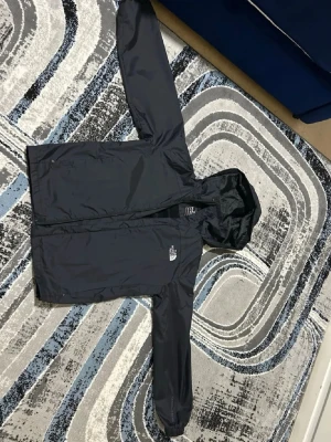 Svart regnjacka från The North Face - Snygg svart regnjacka från The North Face med huva och dragkedja framtill. Jackan har två sidofickor och är tillverkad i ett lätt, vattenavvisande material. Perfekt för regniga dagar och enkel att bära med sig. (Pris kan diskuteras)