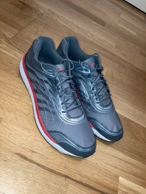 Sportiga skor - Säljer ett par nya grå sportiga skor med röda detaljer. Skorna har grå snören och svart/vit sula. Skorna är helt nya men saknae prislapp och förpackning. Perfekta för sport och träning, men funkar också som vardagliga skor.