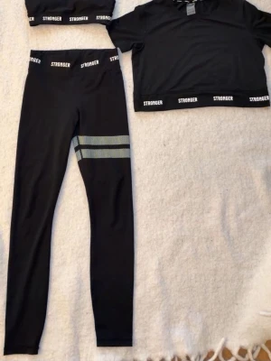Svart träningsset från Stronger - Snyggt svart träningsset från Stronger med sport-bh, croppad t-shirt och tights. Tightsen har två ljusgröna ränder på ena benet och logga på resåren. Materialet är stretchigt och består till stor del av återvunnet tyg. Perfekt för gym eller löpning. Byxorna-s   bh-xs   Topp-xs