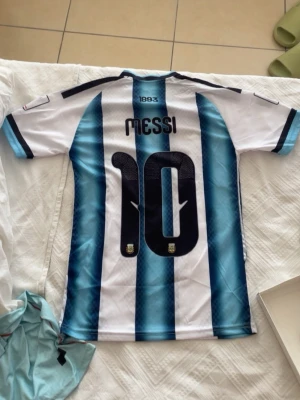 Argentina Messi matchtröja blåvit - Säljer en officiell Argentina matchtröja med Messi och nummer 10 på ryggen. Tröjan har klassiska blå och vita ränder, korta ärmar och detaljer i svart och guld. Perfekt för fotbollsfans som vill stötta sitt lag med stil.